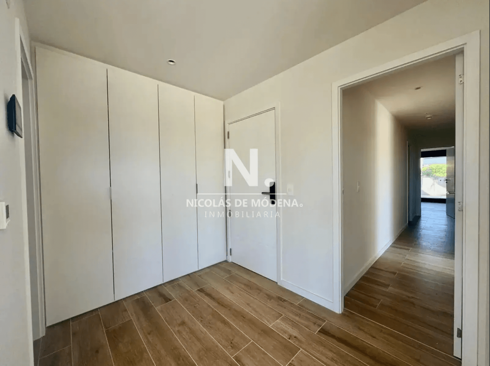 This image captures a bright, modern interior space, likely a hallway or dressing area, featuring extensive built-in white wardrobe storage along one wall. The flooring consists of warm-toned, wood-look rectangular tiles laid in a running bond pattern. An open doorway leads into a brightly lit corridor, suggesting access to other rooms or an exterior space.