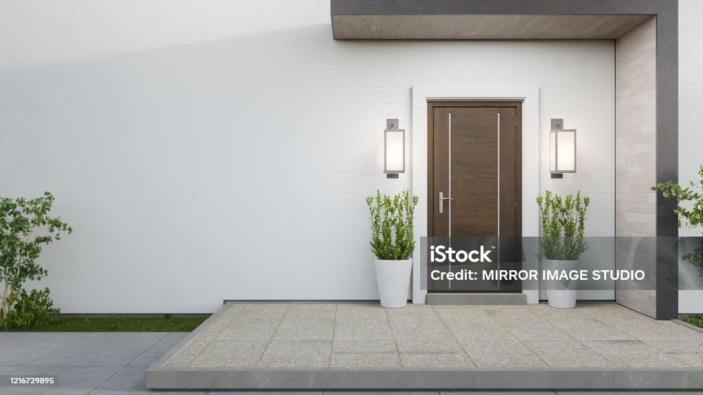 This image features the exterior entrance of a modern home, showcasing a clean, minimalist design. A dark wood front door is flanked by contemporary wall sconces and two potted green plants, set against a white stucco wall. The entrance is accessed via a light-colored tiled patio with a raised concrete edge, leading to a small patch of grass and a shrub on the left.
WATERMARK: iStock