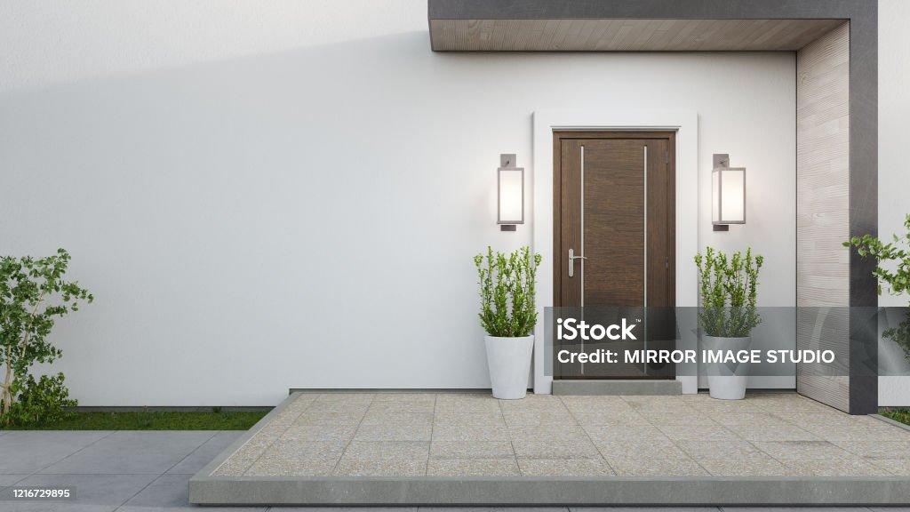 This image features the exterior entrance of a modern home, showcasing a clean, minimalist design. A dark wood front door is flanked by contemporary wall sconces and two potted green plants, set against a white stucco wall. The entrance is accessed via a light-colored tiled patio with a raised concrete edge, leading to a small patch of grass and a shrub on the left.
WATERMARK: iStock