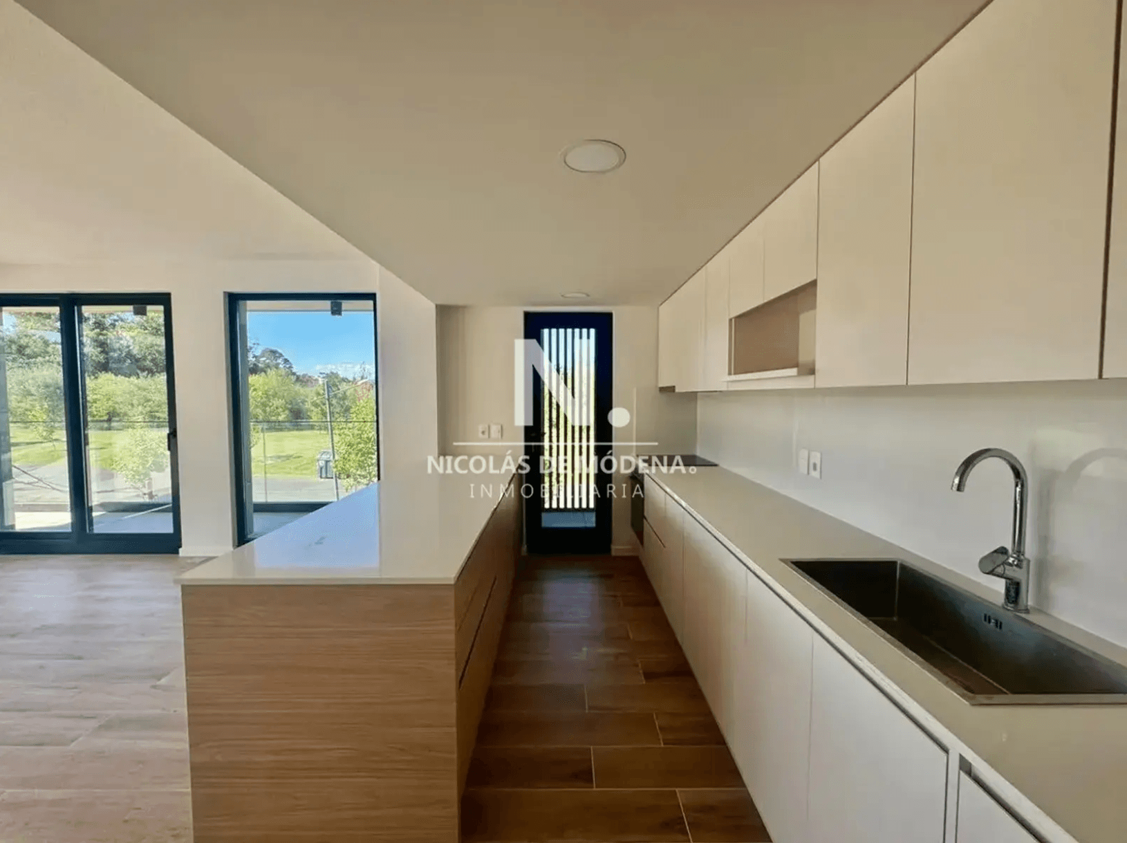 This modern kitchen features sleek white cabinetry and countertops, with a stainless steel sink and a contemporary faucet. A light wood-toned island with a white countertop provides additional workspace, while large glass doors offer views of the exterior greenery.
WATERMARK: NICOLÁS DE MÓDENA. INMOBILIARIA