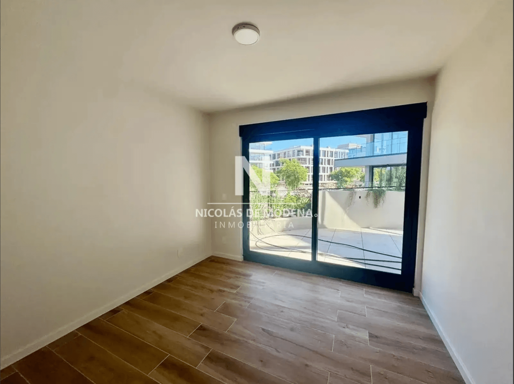 This is a bright, unfurnished room featuring light wood-look tile flooring and white walls. A large sliding glass door with a dark frame opens to an outdoor patio area, offering views of modern buildings and greenery. A simple circular ceiling light fixture is present.
WATERMARK: NICOLÁS DE MODENA INMOBILIARIA
