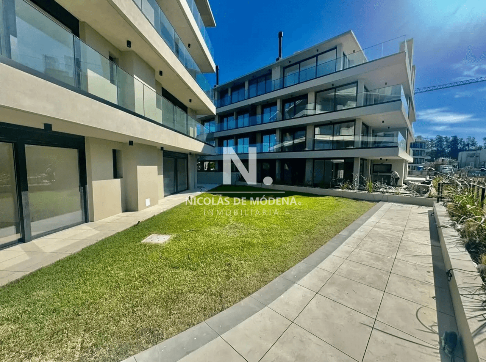 Exterior view of a modern apartment building featuring multiple levels with glass balconies and large windows. The ground floor units have direct access to a well-maintained lawn and a tiled walkway.
WATERMARK: Nicolás de Módena Inmobiliaria