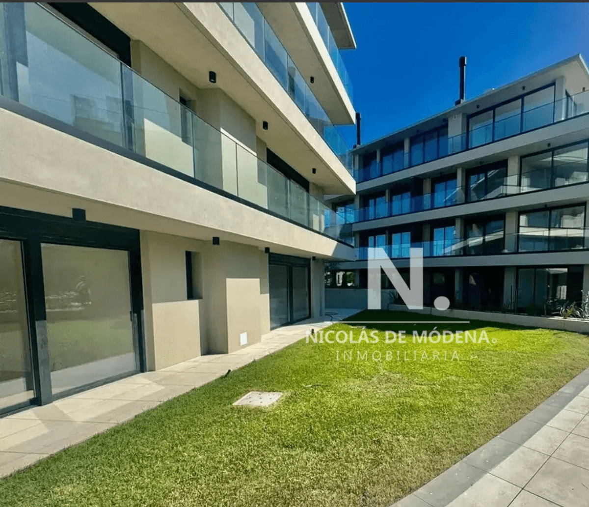 Exterior view of a modern apartment complex featuring two multi-story buildings with light-colored facades and extensive glass balconies. A well-maintained green lawn separates the ground-floor units, which have large sliding glass doors. The property appears new or recently renovated under a clear blue sky.
WATERMARK: Nicolás de Módena Inmobiliaria
