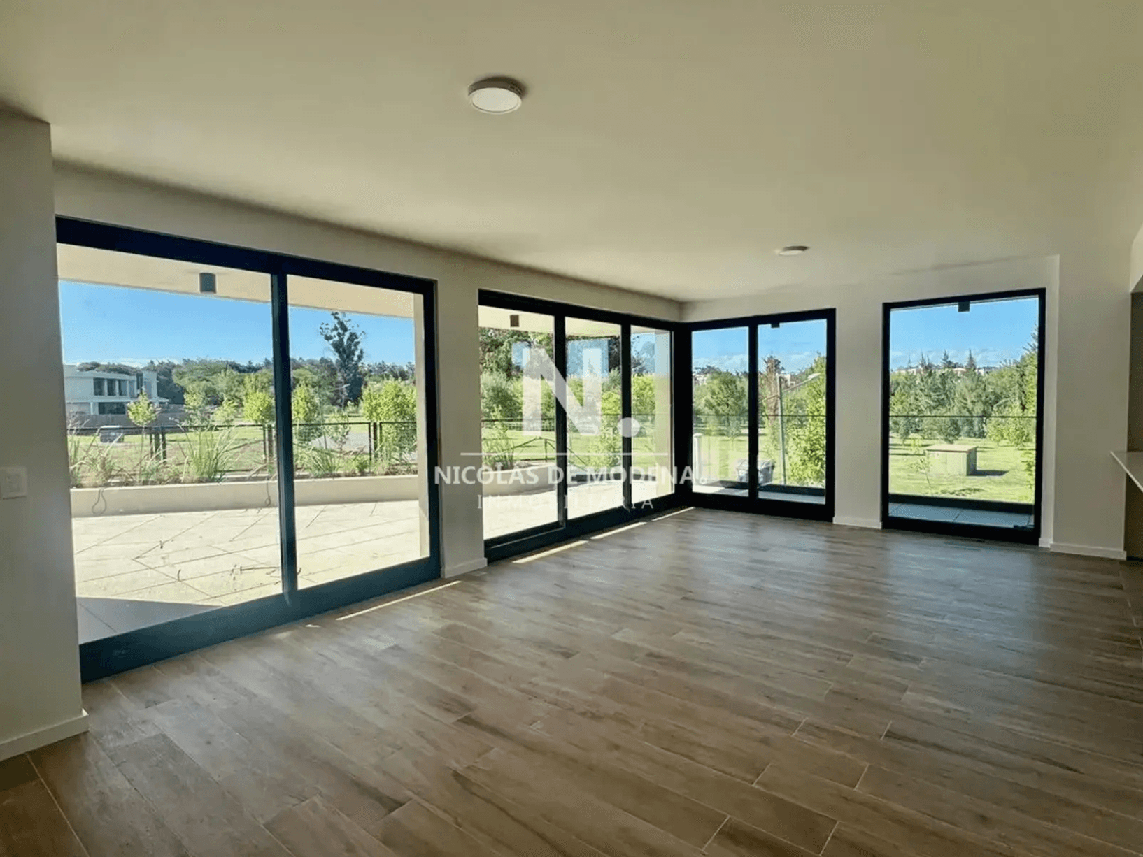 This is a spacious living area featuring light-colored walls and modern wood-look tile flooring. The room is well-lit by multiple large sliding glass doors and windows with black frames, offering extensive views of the outdoor patio and green landscape. Recessed lighting fixtures are visible on the ceiling.
WATERMARK: NICOLAS DE MODENA