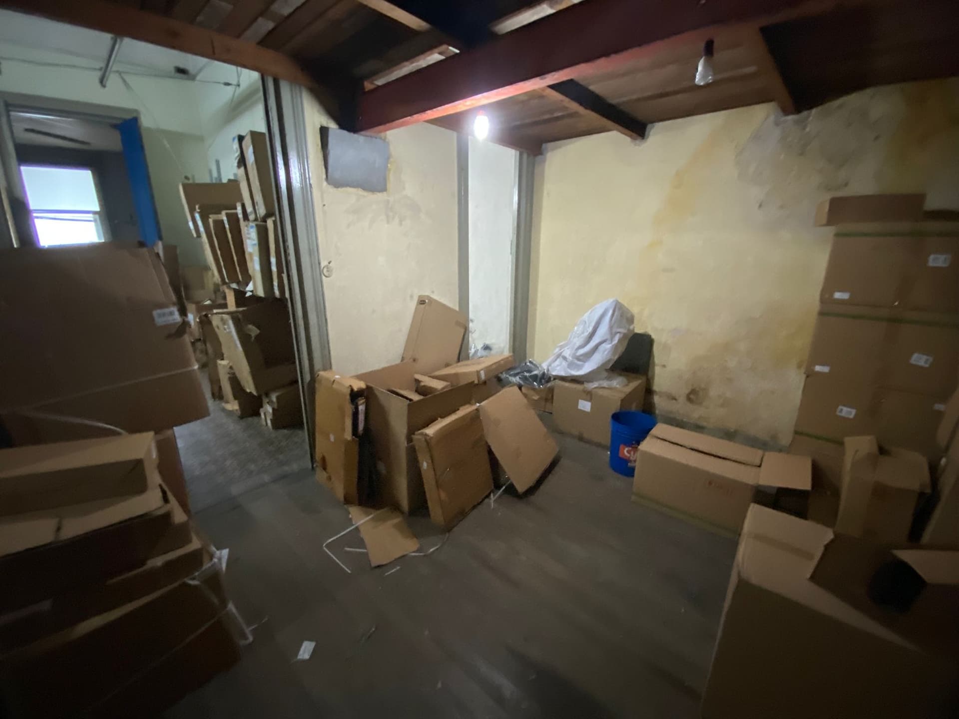 This image depicts an interior storage space or utility room in poor condition. The room features exposed wooden ceiling beams, discolored and damaged walls, and a concrete floor. Numerous cardboard boxes are stacked throughout the space, indicating its use for storage.