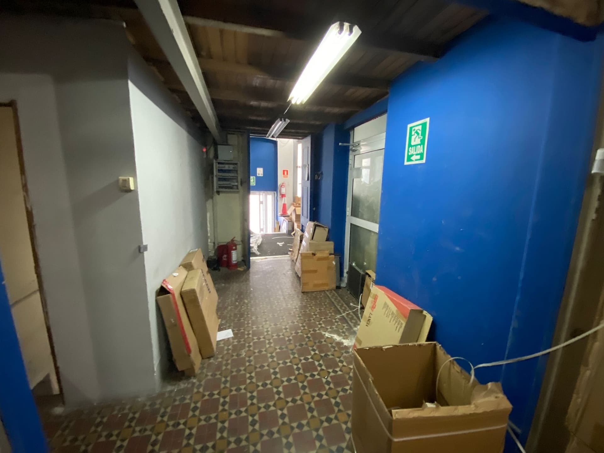 This image captures an interior hallway or corridor, currently used for storage, featuring a patterned tile floor and a wooden plank ceiling. The space is illuminated by fluorescent strip lighting, and includes a blue wall with an "EXIT" sign, a fire extinguisher, and various cardboard boxes. The overall condition appears to be functional but cluttered, suggesting a commercial or industrial setting.