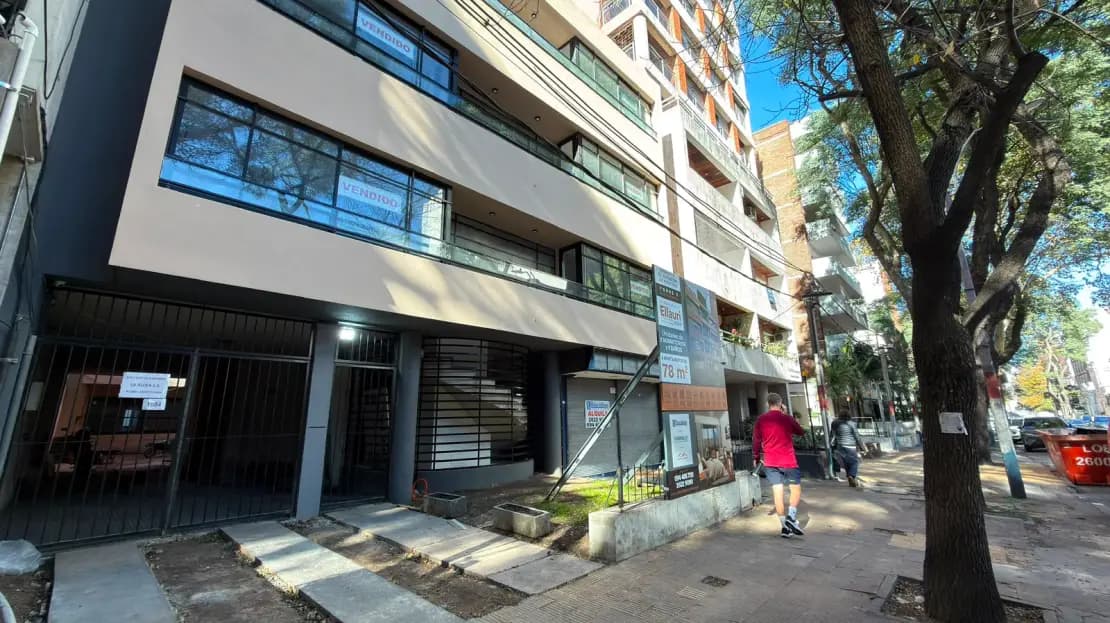 # Property Exterior - Mixed-Use Building
This image shows the street-level facade of a modern multi-story residential or mixed-use building featuring clean white and dark gray finishes with contemporary horizontal lines. The ground floor includes commercial storefronts with black metal security gates and large windows, while upper floors display residential units with balconies and modern glazing. The building is situated on an active urban street lined with mature trees, with good natural lighting and pedestrian accessibility.