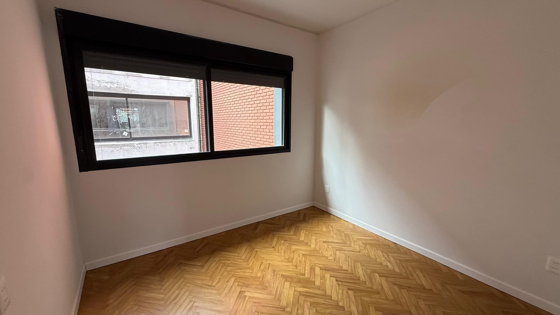 # Property Image Description
This is a bright, modern bedroom or living space featuring clean, neutral beige walls and herringbone-pattern wooden flooring in warm honey tones. The room is well-lit by a large horizontal window with a sleek black frame that offers views of the exterior brick building facade. The space appears freshly renovated with excellent natural light and minimal furnishings, showcasing the room's generous proportions and contemporary finishes.