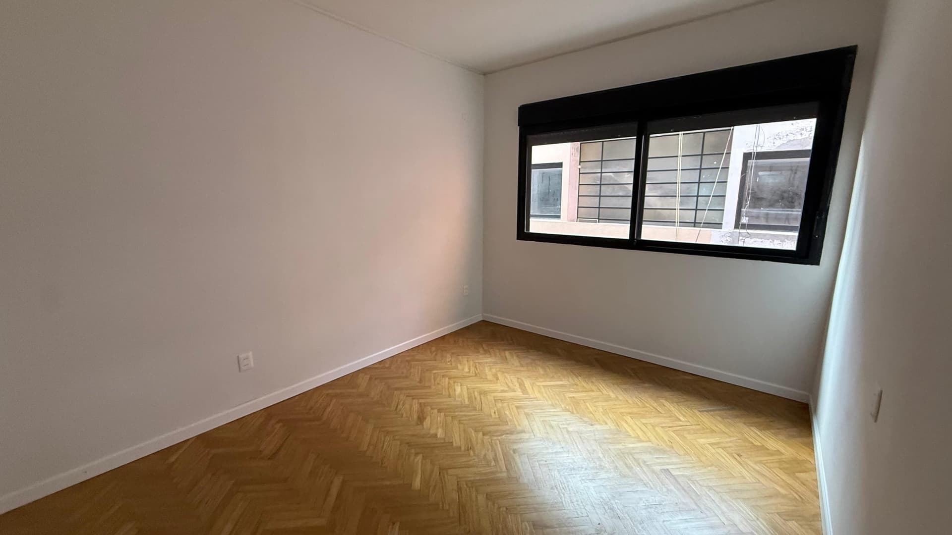 This is a bright, unfurnished bedroom or living space featuring clean white walls and herringbone-pattern wooden flooring in good condition. The room is well-lit by a large black-framed sliding window that provides views of the street and neighboring buildings, offering natural light and ventilation. The space is empty and move-in ready, with visible electrical outlets on the walls.