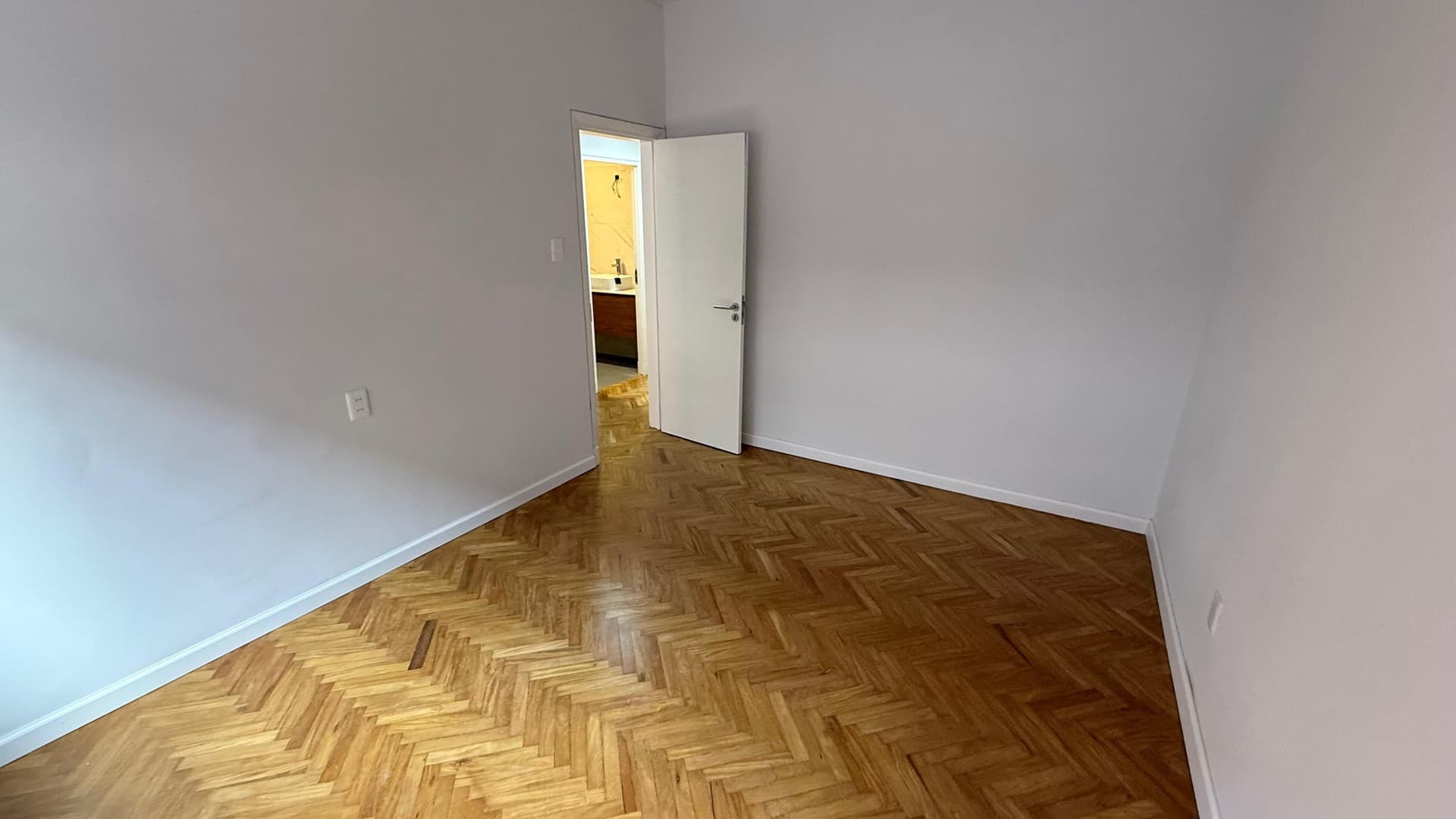 # Bedroom
This spacious bedroom features classic herringbone parquet flooring in excellent condition and clean white walls with fresh paint. The room includes a white interior door providing access to an ensuite bathroom with yellow tile accents, and benefits from bright natural lighting throughout the space.