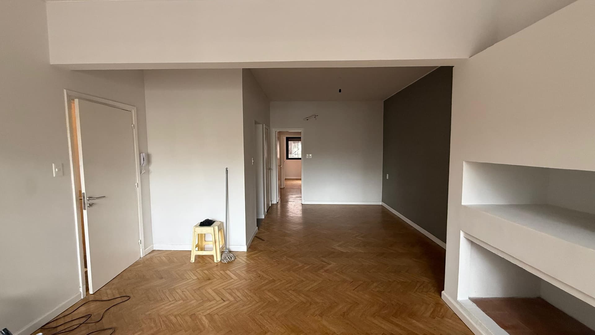 # Property Image Description
This is a spacious open-plan living area or apartment interior featuring herringbone-pattern hardwood flooring throughout. The space showcases clean white walls with modern architectural elements, including built-in shelving on the right side and a distinctive dark accent wall in the background. Multiple doorways provide access to adjoining rooms, and the area benefits from natural light streaming through windows visible in the distance, creating a bright and airy atmosphere. The property appears to be under renovation or preparation, with minimal furnishings present.