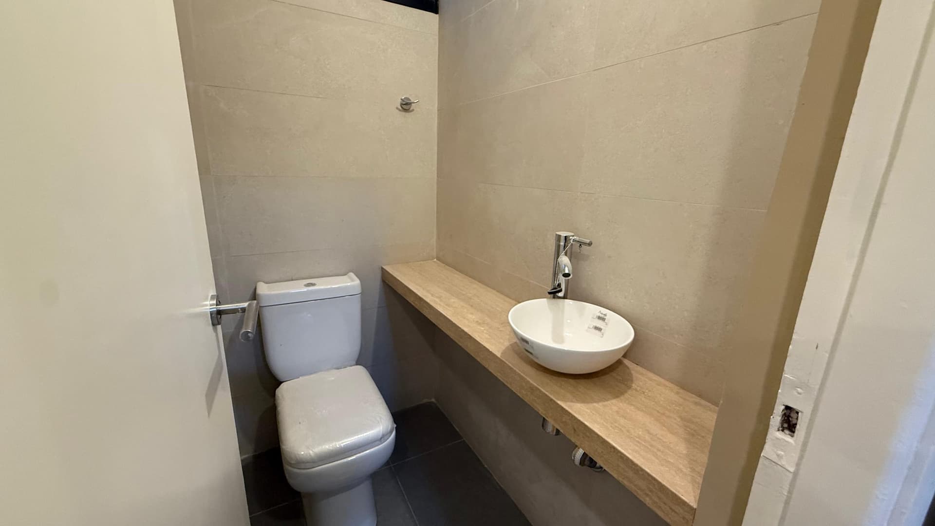 # Bathroom Description
This is a modern, compact bathroom featuring a white ceramic toilet on the left and a contemporary round vessel sink mounted on a natural wood countertop with a chrome faucet. The space is finished with neutral beige tile walls and dark flooring, creating a clean, minimalist aesthetic with updated fixtures and contemporary design elements.