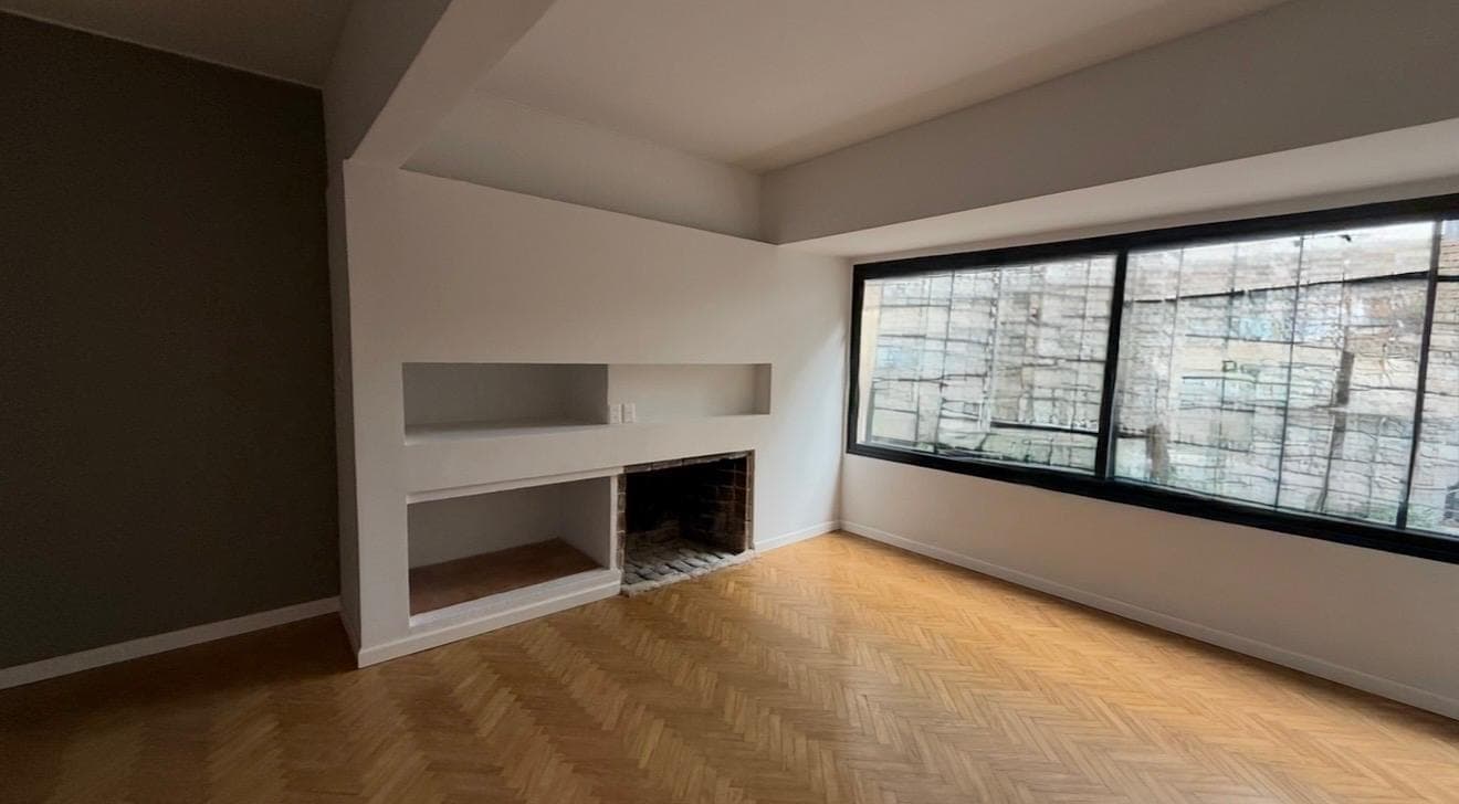 # Living Room with Fireplace
This spacious living room features a modern fireplace with built-in shelving and a clean white surround, complemented by herringbone-pattern hardwood flooring in warm honey tones. Large floor-to-ceiling windows with black frames provide abundant natural light and city views, while the room benefits from contemporary architectural details including a coffered ceiling and neutral color palette with an accent dark wall.