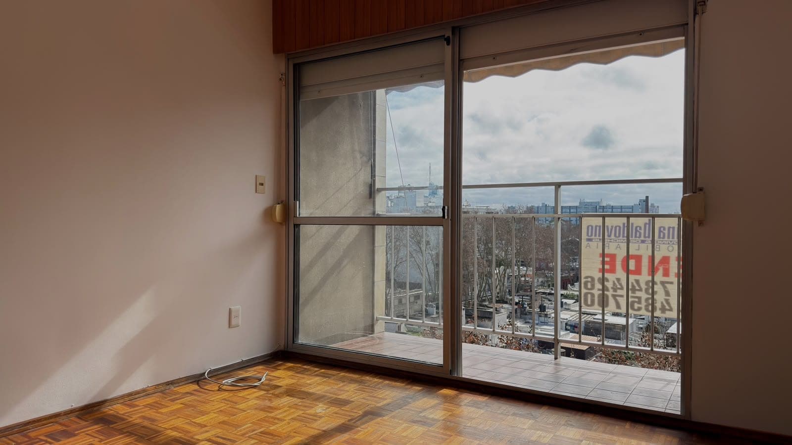 # Property Image Description
This is a high-rise residential space featuring a spacious room with expansive floor-to-ceiling sliding glass doors that open onto a balcony with panoramic city views. The room showcases warm wood parquet flooring, soft pink/mauve walls, and dark wood cabinetry above. The balcony offers sweeping vistas of an urban skyline with distant waterfront views, and includes a retractable awning for shade. The space is bright and airy with excellent natural light throughout.