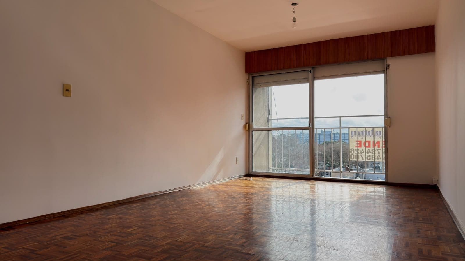 # Property Image Description
This is a spacious living room or bedroom featuring herringbone wood flooring in excellent condition and neutral beige walls. The room is flooded with natural light from expansive sliding glass doors that open to a balcony with panoramic city views, complemented by burgundy fabric window treatments. The space is clean and move-in ready, with modern fixtures including a ceiling-mounted light and electrical outlets visible on the walls.
