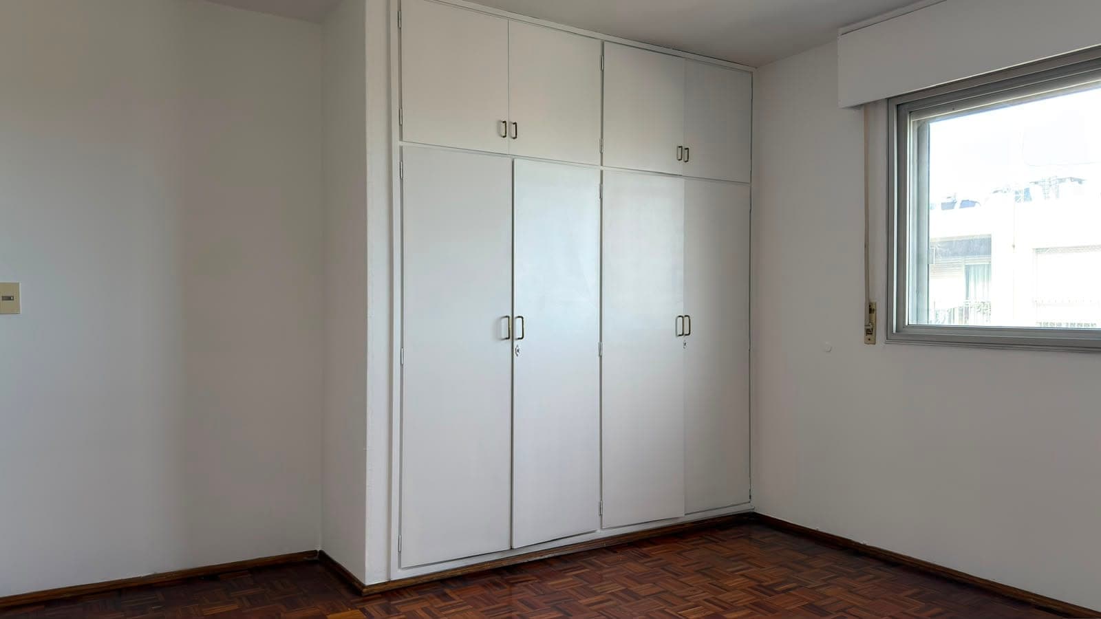 # Bedroom with Built-in Wardrobes
This bright bedroom features extensive built-in white cabinetry with multiple mirrored wardrobe doors and chrome handles, providing substantial storage solutions. The room has herringbone-pattern wooden flooring, neutral white walls, and a large window with city views, creating a clean and contemporary aesthetic. Natural light floods the space, highlighting the modern finishes and well-maintained condition of this residential bedroom.