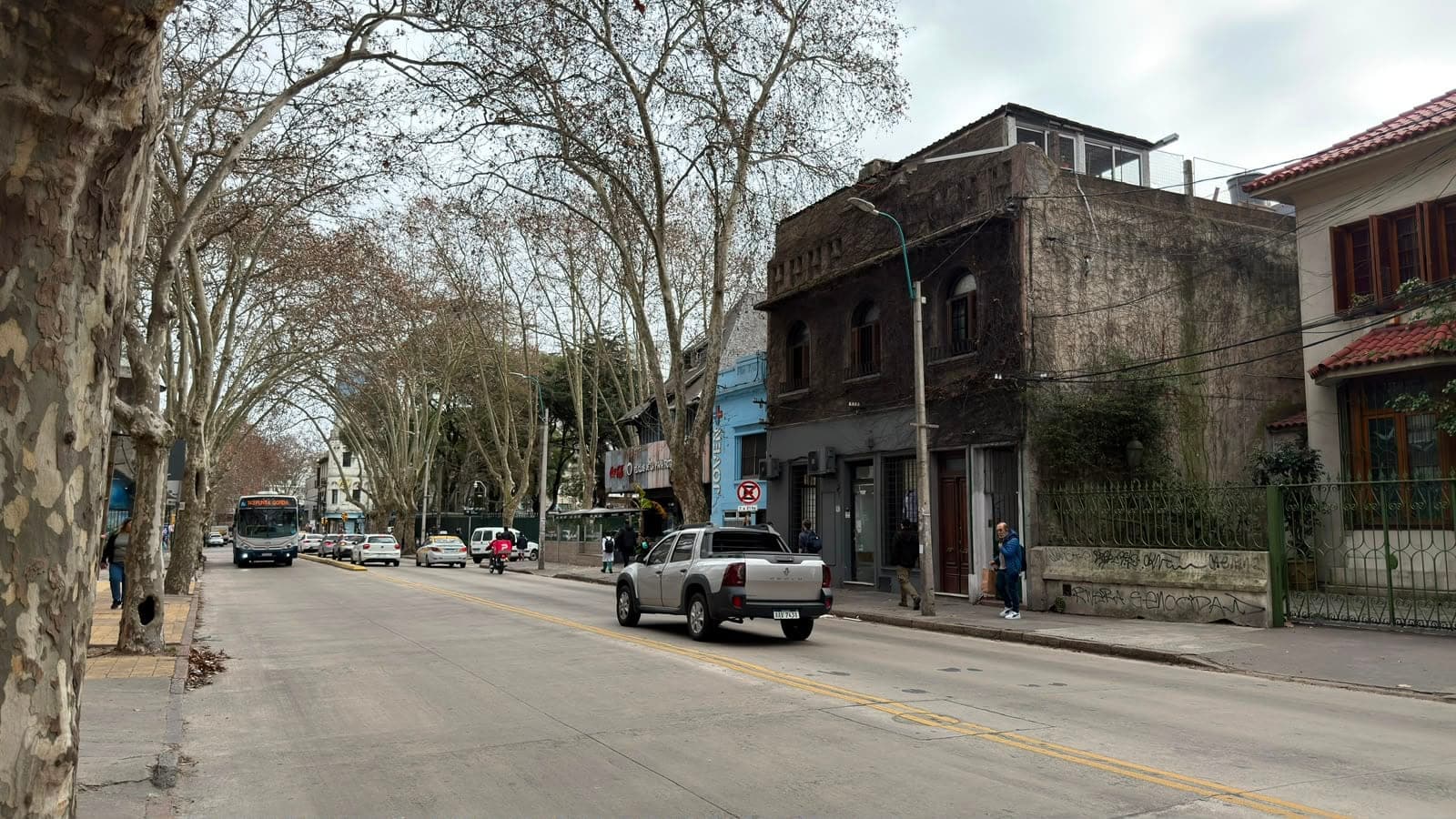 # Street View - Historic Urban Neighborhood
This image shows a tree-lined street in a historic urban district featuring a mix of architectural styles. The focal point is a weathered, multi-story building with dark stone facade and a modern rooftop addition, displaying significant age and patina. Adjacent properties include a bright blue colonial-style building and a cream-colored structure with red tile roofing, creating a diverse streetscape typical of Latin American heritage neighborhoods with mature plane trees providing canopy coverage.