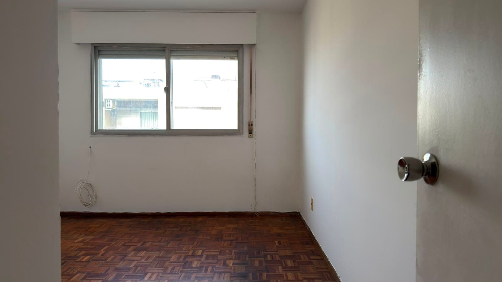 # Property Image Description
This is a bright, unfurnished bedroom or living space featuring clean white walls and a geometric parquet wood floor in warm brown tones. The room is well-lit by a large sliding window with a modern frame that overlooks an urban cityscape with neighboring buildings visible. The space includes basic fixtures such as a door knob on the right wall and electrical outlets, with minimal visible wear, suggesting the property is ready for renovation or move-in.