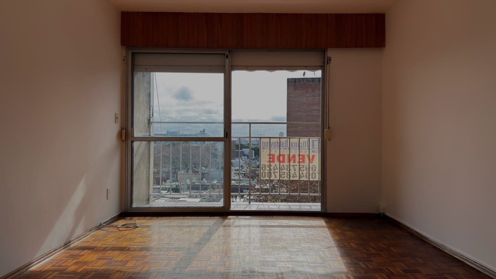 # Property Image Description
This is a spacious high-rise bedroom or living room featuring expansive floor-to-ceiling sliding glass doors that open onto a balcony with panoramic city views. The room showcases hardwood parquet flooring in warm tones and neutral beige walls, with a dark wood valance above the windows. The large windows flood the space with natural light and offer views of an urban skyline with brick buildings and distant cityscape, making this an ideal space for those seeking elevated city living with excellent natural illumination.