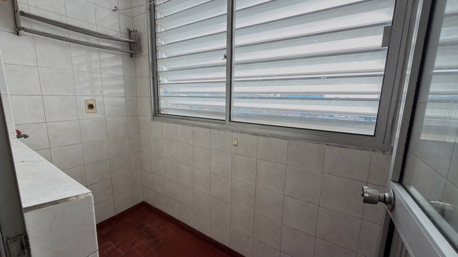 This is a bathroom featuring light gray ceramic tile walls and a contrasting dark red tile floor. The space includes a white bathtub on the left side and large windows with horizontal blinds that provide natural light and views of the surrounding area. The bathroom is in functional condition with basic fixtures and good natural ventilation from the multi-pane windows.