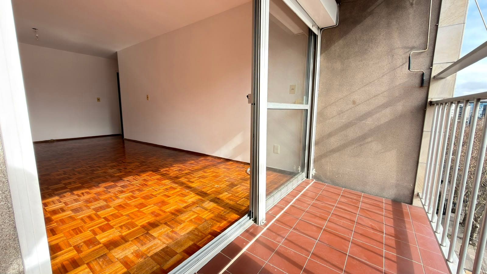 # Property Image Description
This image shows a spacious living room with an adjoining balcony/terrace area. The interior features beautiful warm-toned parquet flooring in a herringbone pattern, white walls, and abundant natural light from large glass sliding doors that open to the outdoor space. The terrace is finished with terracotta-colored ceramic tiles and includes a white metal railing, offering what appears to be an elevated view of the surrounding area.