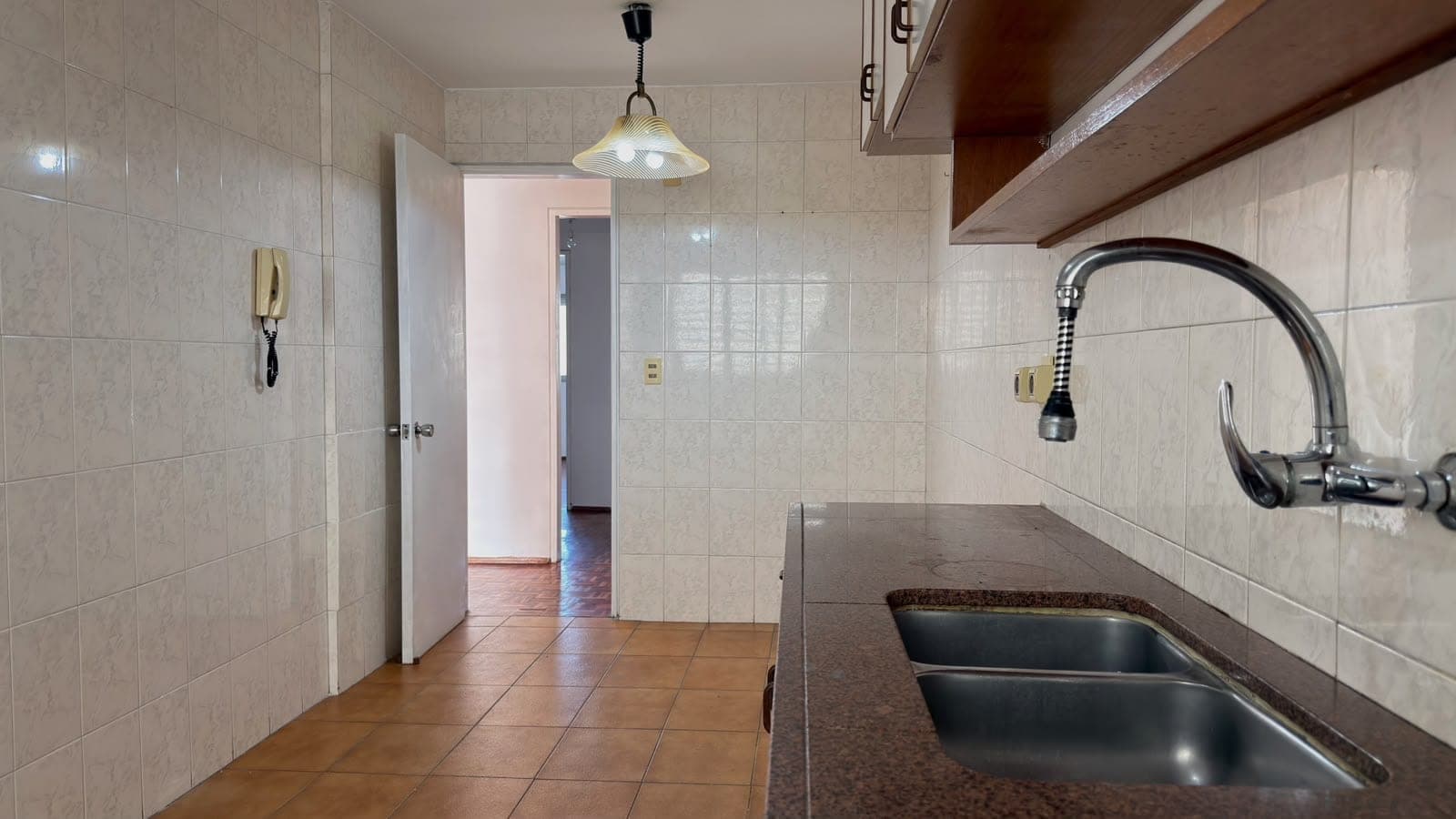 # Kitchen
This galley-style kitchen features cream-colored ceramic tile walls and terracotta tile flooring in good condition. The space includes a dark granite countertop with a stainless steel double sink, pull-down faucet, and open shelving above. A pendant light fixture provides overhead illumination, and the kitchen opens to adjacent living areas through a doorway, offering functional layout and practical storage solutions.
