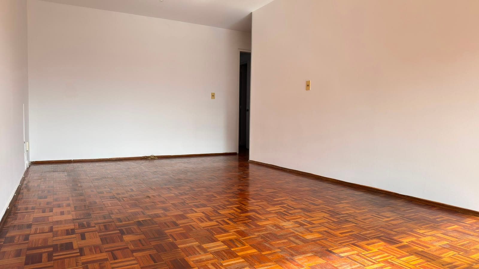 # Property Description
This is a spacious, unfurnished living room or bedroom featuring beautiful hardwood parquet flooring in a warm honey-brown tone with a geometric pattern. The room has clean, neutral beige walls, recessed lighting, and brass electrical outlets, creating a bright and airy atmosphere. A dark wooden door is visible in the background, and the space appears to be in excellent condition, ready for occupancy or customization.