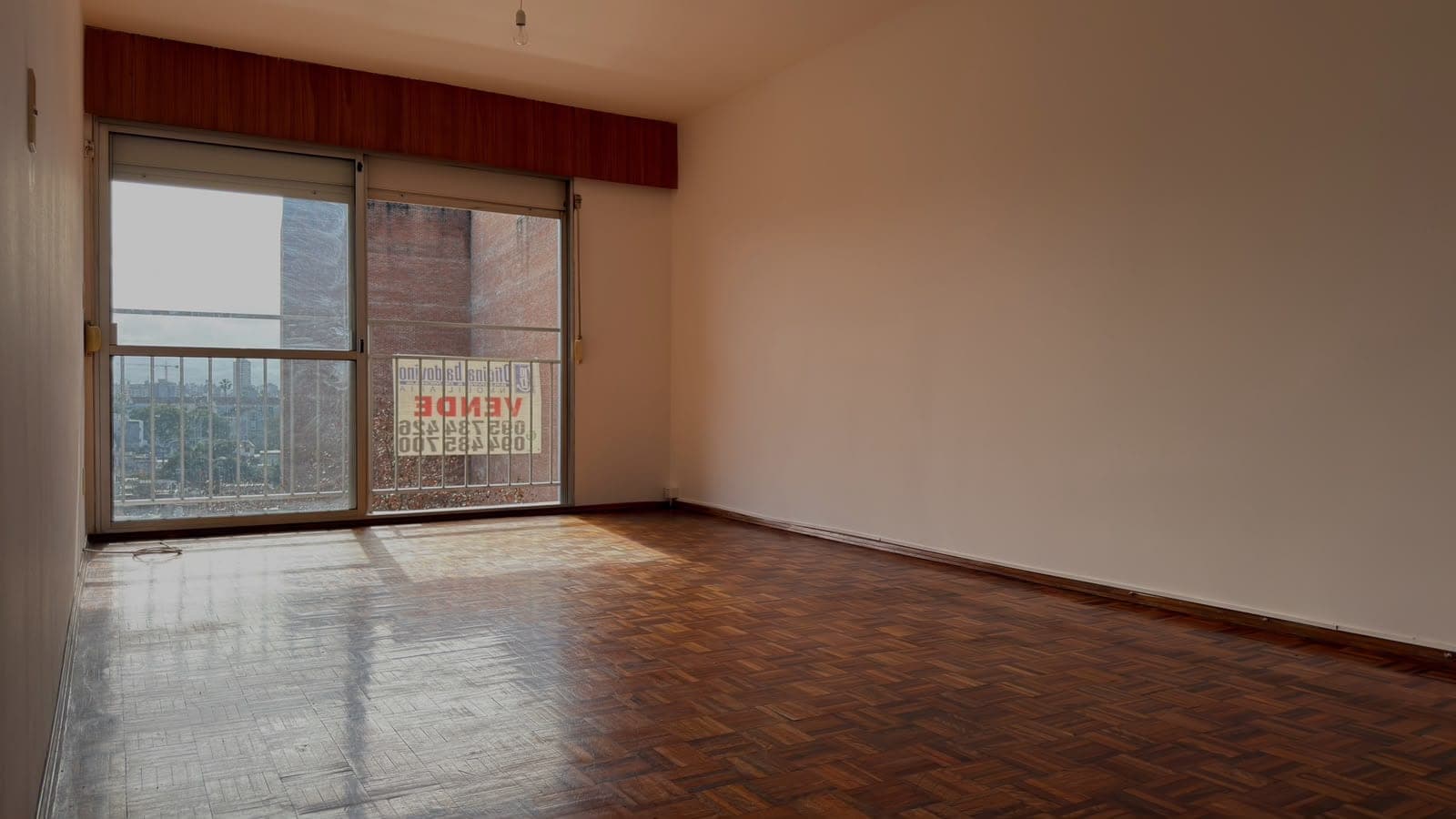 # Property Image Description
This is a spacious living room or bedroom featuring warm peachy-beige walls and herringbone parquet flooring in good condition. The room is flooded with natural light from large sliding glass doors with a burgundy valance, offering expansive city views and access to a balcony overlooking urban architecture. The space is empty and move-in ready, with clean lines and neutral finishes ideal for various interior design styles.