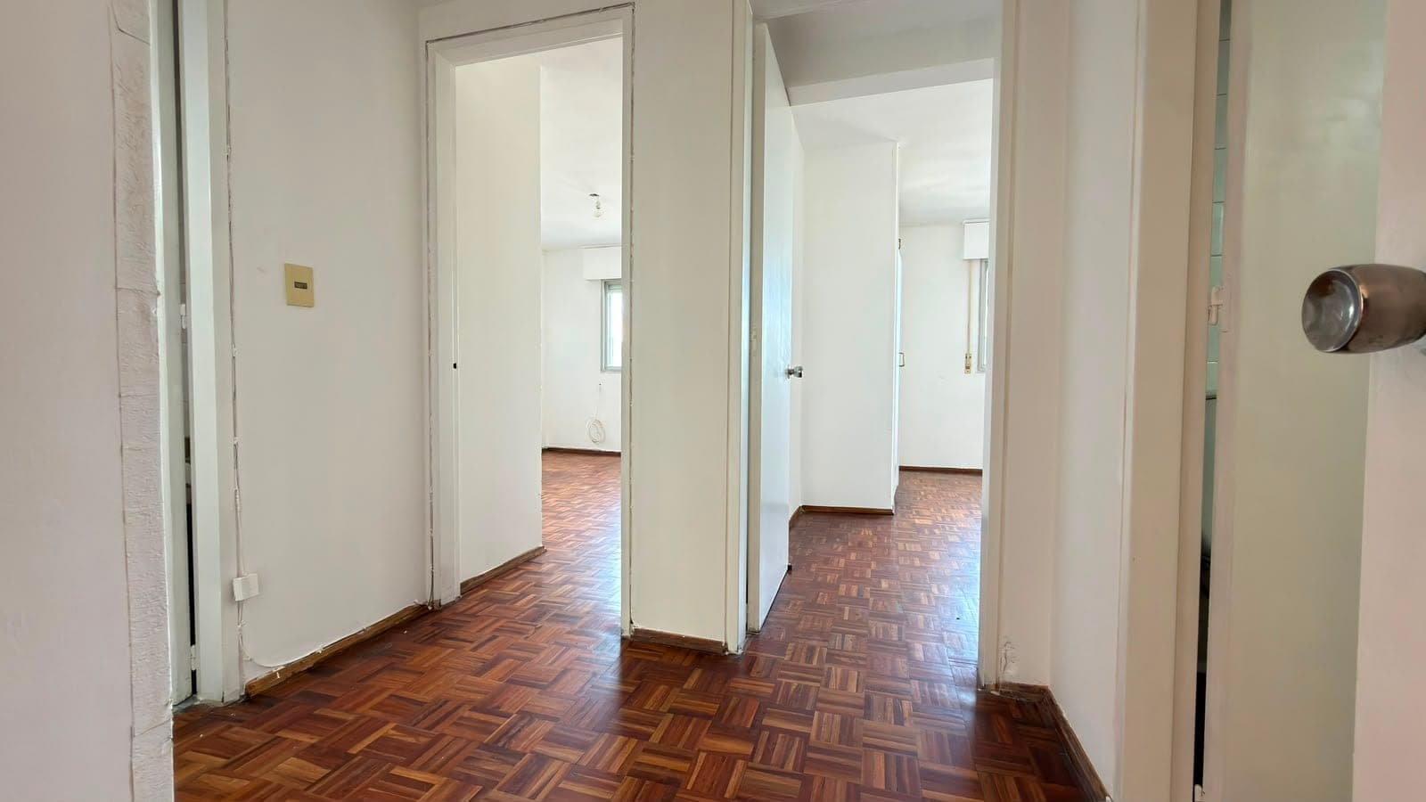 # Property Image Description
This is an interior hallway or corridor featuring classic herringbone parquet flooring in rich wood tones. The space is bright and clean with white painted walls, multiple white interior doors leading to adjacent rooms, and brass/gold-toned electrical fixtures visible on the walls. The hallway demonstrates good natural lighting and provides clear sightlines through multiple doorways, suggesting a well-connected floor plan typical of traditional apartment layouts.