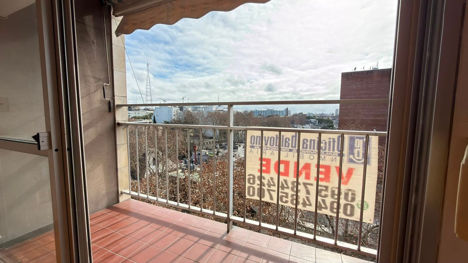 # Property Description
This is a spacious balcony or terrace with terracotta tile flooring and metal railing, offering expansive views of the surrounding cityscape and distant skyline. The space features a retractable brown awning for shade and direct access from an interior room via glass sliding doors. The elevated vantage point provides panoramic views of residential and commercial buildings, with clear sightlines across the urban landscape under partly cloudy skies.
