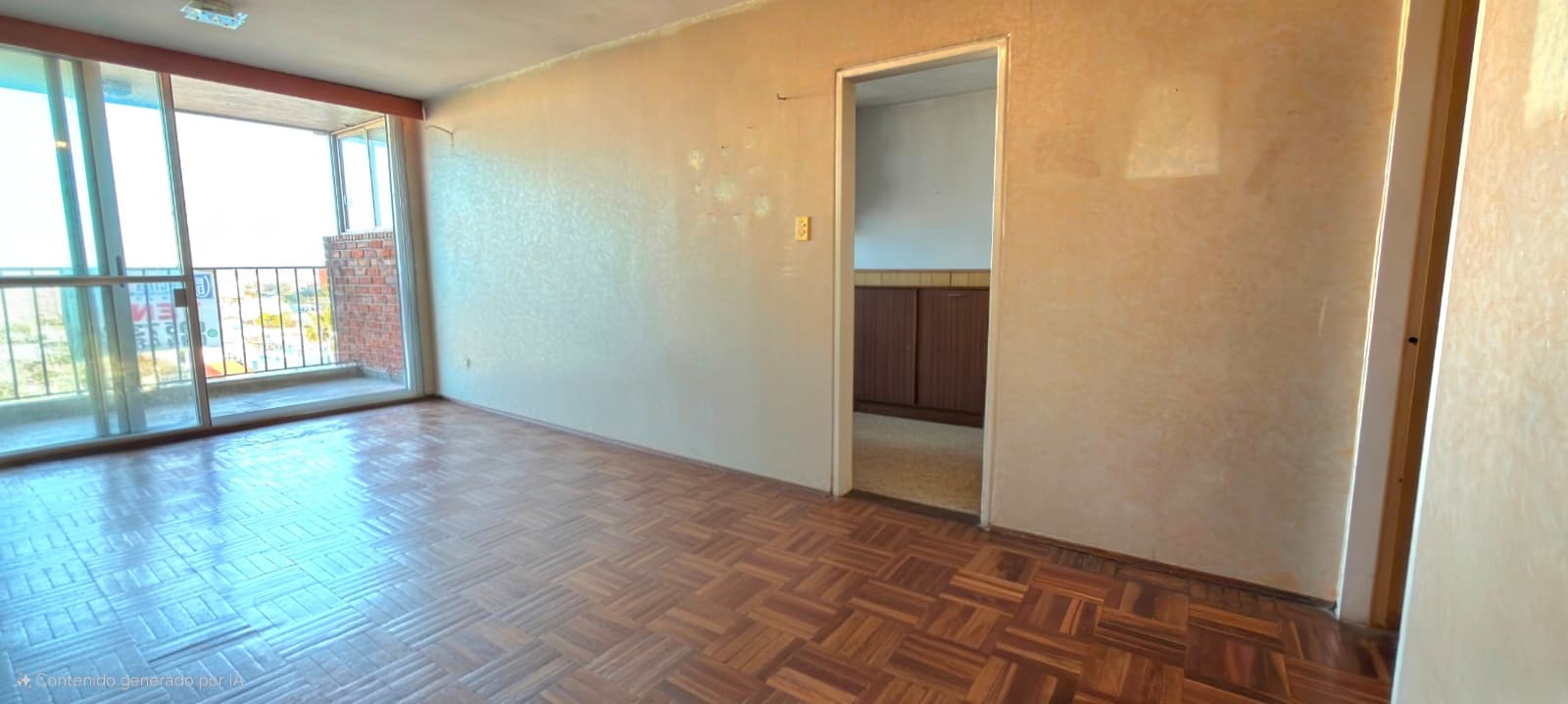 # Property Description
This is a spacious living room or main living area featuring herringbone parquet flooring in warm wood tones and neutral beige walls. The room benefits from abundant natural light through large glass sliding doors that open onto a balcony with street views, and includes a separate doorway leading to an adjacent room. The space appears clean and ready for renovation or occupancy, with recessed ceiling lighting and basic electrical outlets visible.
WATERMARK: Contenido generado por IA