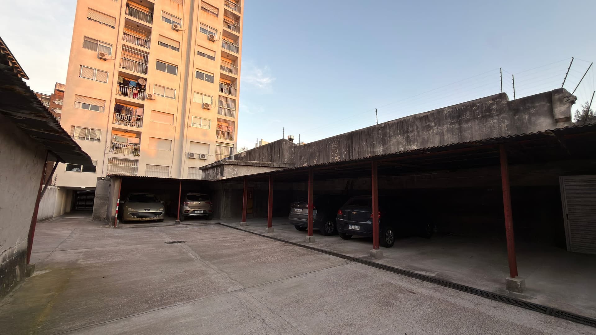 # Property Description
**Covered Parking Area/Carport** - This is an open-air covered parking structure with multiple vehicle spaces supported by red metal columns. The facility shows weathered concrete flooring and a corrugated metal roof with visible age and wear. Currently accommodating several parked vehicles, the carport is adjacent to a multi-story residential apartment building with peach-colored facade, indicating this is part of a larger residential complex.