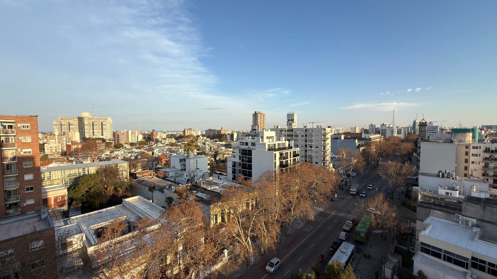 # Elevated Urban View
This image captures an expansive cityscape view from a high-rise vantage point, showcasing a dense urban residential neighborhood with a mix of mid-rise apartment buildings, older brick structures, and modern white residential towers. The scene features clear blue skies, abundant natural light during golden hour, and a sprawling panoramic vista extending across the city skyline with communication towers visible in the distance. The foreground displays deciduous trees in late autumn condition and street-level activity, suggesting this view is from a upper-floor apartment or balcony in an established urban area.