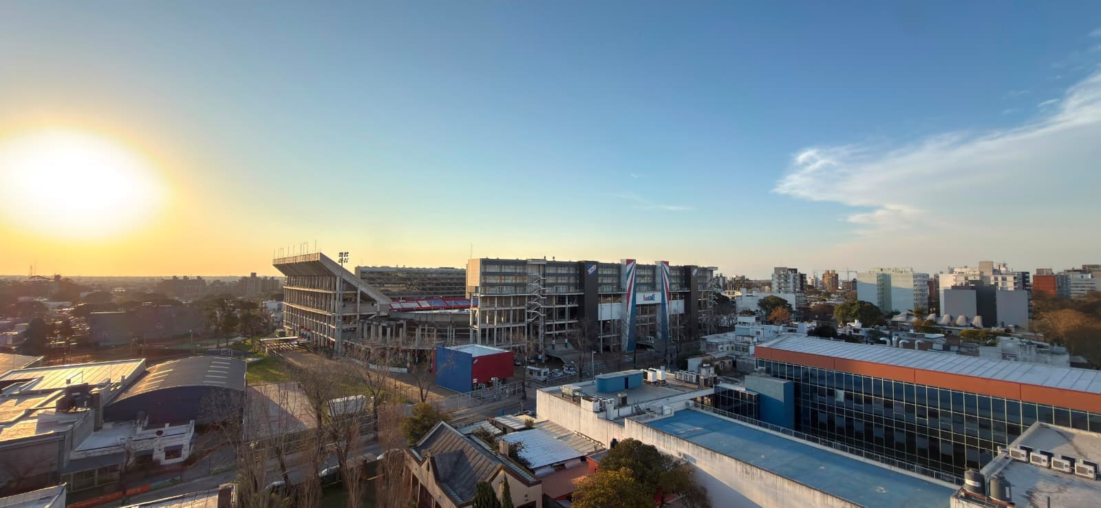 # Property Image Description
This is an elevated exterior view capturing a vibrant urban mixed-use development during golden hour. The scene showcases modern residential and commercial buildings with contemporary architecture, including a prominent sports stadium, contemporary apartment complexes with dark facades, and a blue-roofed recreational facility in the foreground. The expansive cityscape view extends across the skyline with clear visibility of surrounding neighborhoods, green spaces, and distant mountains, offering excellent natural light and panoramic vistas from this elevated vantage point.