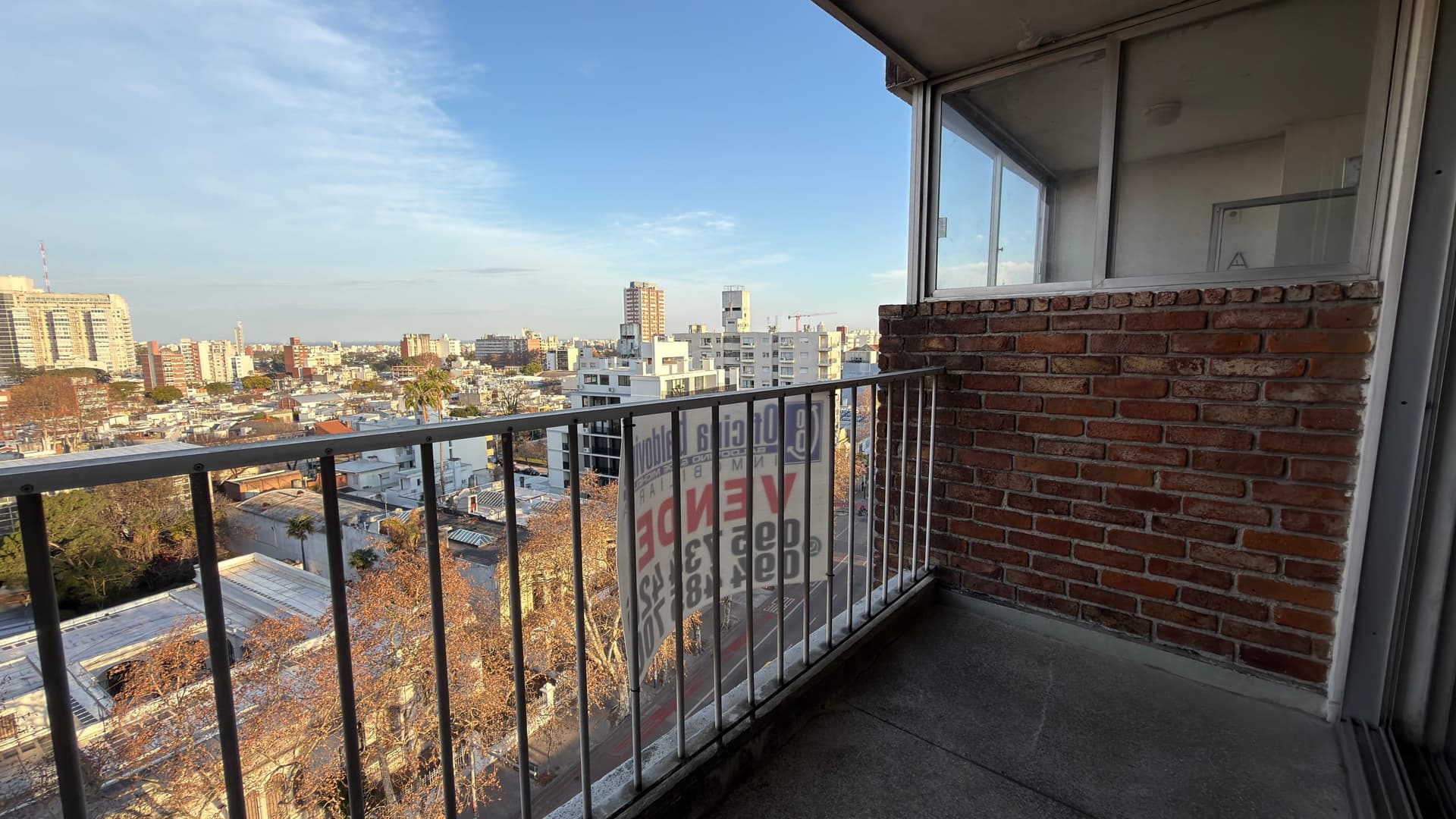 # Property Description
This is a high-rise balcony/terrace space offering expansive panoramic city views of an urban skyline with multiple residential and commercial buildings visible in the distance. The balcony features modern black metal railings, a concrete floor, and an exposed red brick accent wall, with large sliding glass doors providing access to the interior living space. The property benefits from excellent natural light and unobstructed views across the cityscape, ideal for a contemporary urban residence.