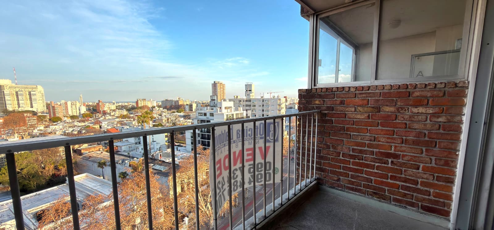 # Property Description
This is a high-rise balcony/terrace space featuring expansive panoramic city views of an urban skyline with multiple residential and commercial buildings. The balcony is equipped with modern black metal railings and large sliding glass doors providing access to the interior, with exposed red brick detailing on the adjacent wall. The space offers excellent natural light and unobstructed vistas across the cityscape under clear blue skies, making it an ideal outdoor entertaining area for an upper-floor residential unit.