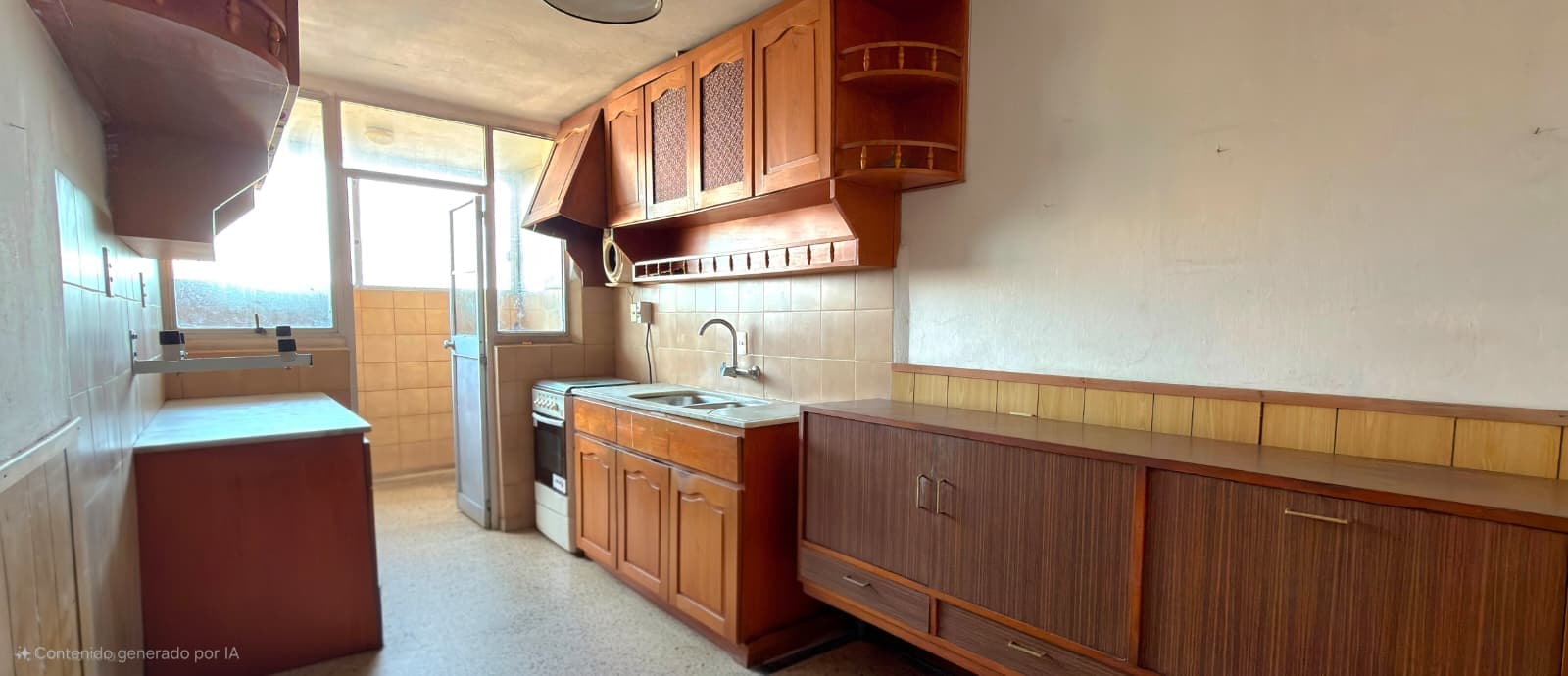 # Kitchen
This is a galley-style kitchen featuring warm wood cabinetry in medium to dark tones with glass-front upper cabinets and textured lower cabinet doors. The space includes a gas stove, sink with faucet, and beige tile backsplash, with ample counter space and storage throughout. Large windows provide natural light and views to the exterior, and the kitchen opens to an adjacent bathroom area visible on the left side of the image.