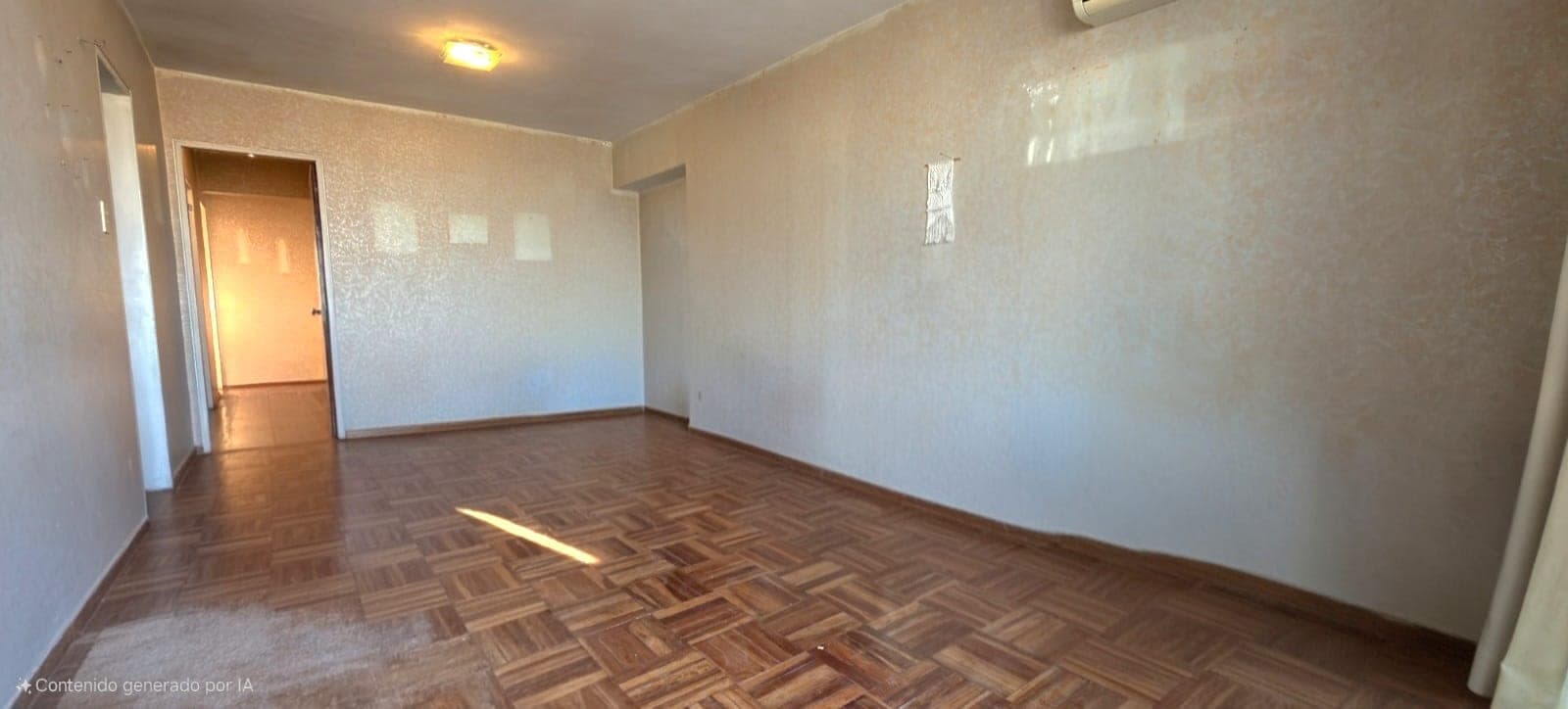 # Property Description
This is a spacious bedroom or living room featuring light beige/cream textured wallpaper and a herringbone-pattern wooden floor in warm brown tones. The room is well-lit with natural sunlight streaming through a window and includes ceiling-mounted lighting fixtures, with visible HVAC vents and electrical outlets on the walls. An open doorway on the left provides access to an adjacent room, and the space appears clean and ready for occupancy.