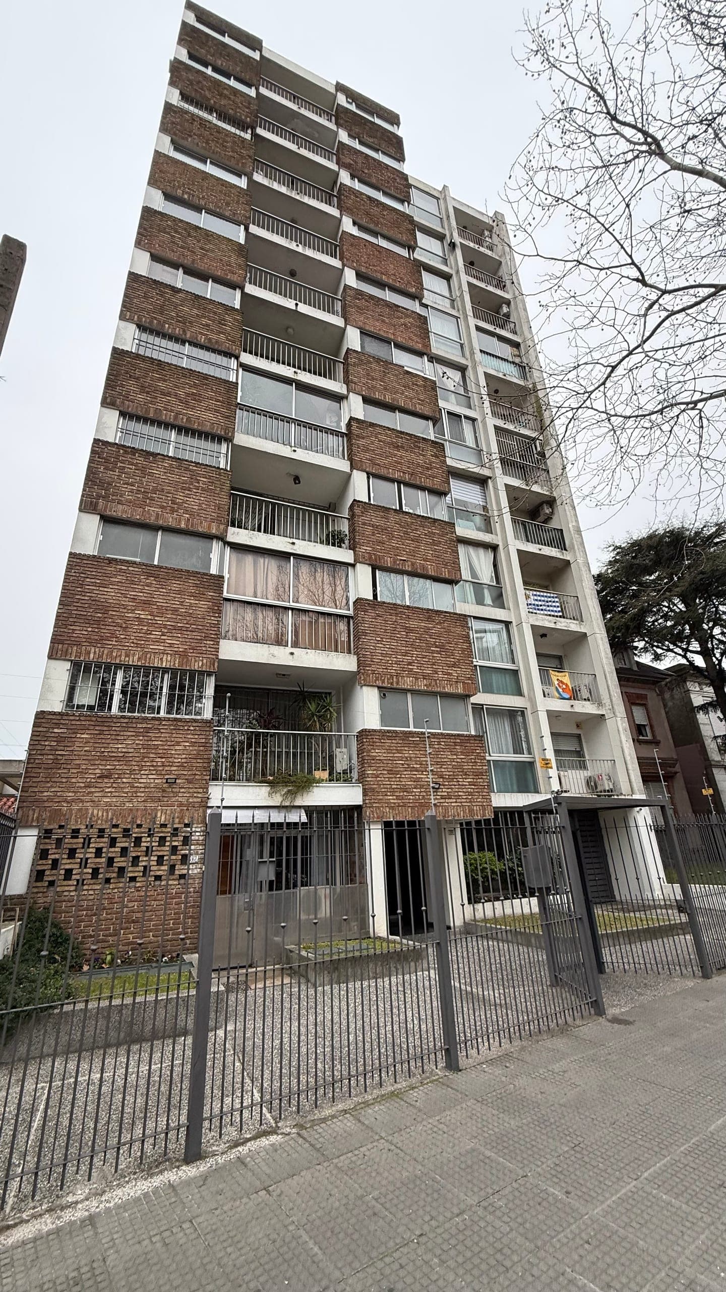 # Property Exterior - Mid-Rise Apartment Building
This is a mid-rise residential apartment building featuring a distinctive brutalist design with alternating bands of red brick and white concrete panels. The structure spans approximately 10 stories and includes individual balconies on each unit, with a secure gated entrance at street level and ground-floor commercial or residential spaces. The building shows typical urban European architecture with well-maintained brick facades, though some weathering is visible on the concrete elements and window frames.