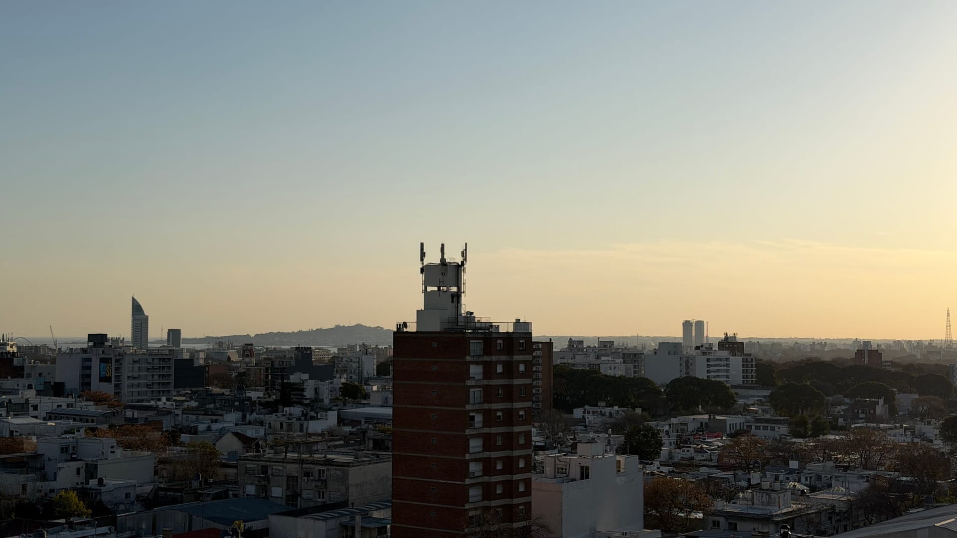 This is an exterior view showcasing a panoramic cityscape from an elevated vantage point, likely from a high-rise residential building. The image captures a sprawling urban landscape with mixed-height buildings, including a distinctive red-brick tower in the foreground with communication antennas, surrounded by modern apartment complexes and residential structures. The distant view reveals mountains on the horizon and clear skies with warm golden-hour lighting, suggesting excellent visibility and a desirable location with expansive views.