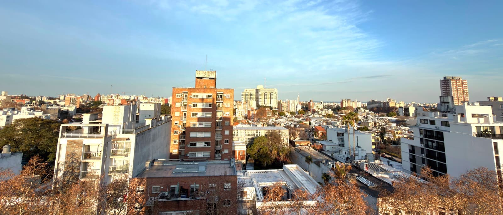 # Elevated Urban Cityscape View
This image captures an expansive cityscape view from an elevated vantage point, showcasing a dense urban neighborhood with mixed architectural styles including mid-rise residential buildings, modern apartment complexes, and older brick structures. The panoramic vista reveals a sprawling metropolitan area with clear blue skies, scattered trees in autumn condition, and diverse building heights creating an interesting skyline. The perspective suggests this view is from a high-floor apartment or rooftop, offering excellent sightlines across the city with multiple neighborhoods visible in the distance.