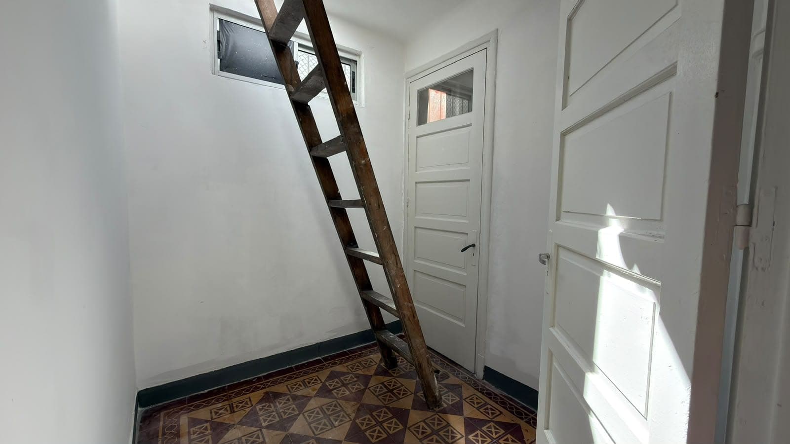 This is an interior hallway or entryway featuring white painted walls and a decorative geometric tile floor in earth tones. The space includes a wooden ladder providing access to an upper level or loft, along with multiple white paneled doors and a small transom window allowing natural light. The room is bright and clean with good natural illumination from the right side, showcasing the original architectural character of what appears to be a historic or period property.