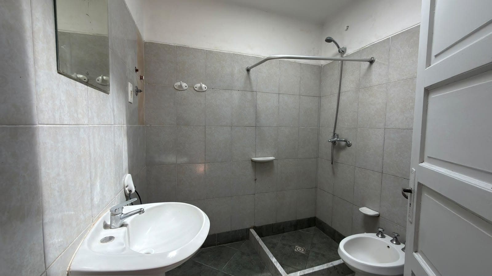 # Bathroom
This is a functional bathroom featuring gray ceramic tile walls and dark tile flooring throughout. The space includes a round white pedestal sink on the left, a walk-in shower area with chrome fixtures and a curved shower rod, a separate small circular sink on the right, and a recessed mirror cabinet above the main sink. The bathroom is clean and well-lit with practical fixtures, though it shows signs of age in the tile finish and overall wear.