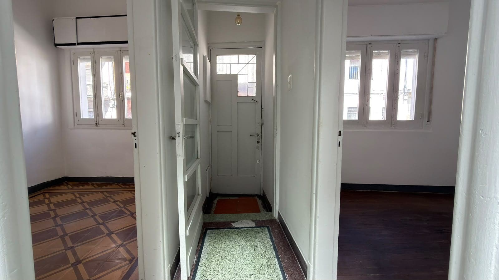 # Property Image Description
**Central Hallway/Entryway**: This bright, well-lit corridor features white walls and multiple doorways leading to adjacent rooms on both sides. The space showcases a mix of flooring types, including geometric patterned tile in the hallway and varied floor finishes in the adjoining rooms. Multiple windows with white frames provide excellent natural light throughout, and the area appears clean and well-maintained with classic architectural details including paneled doors and trim work.