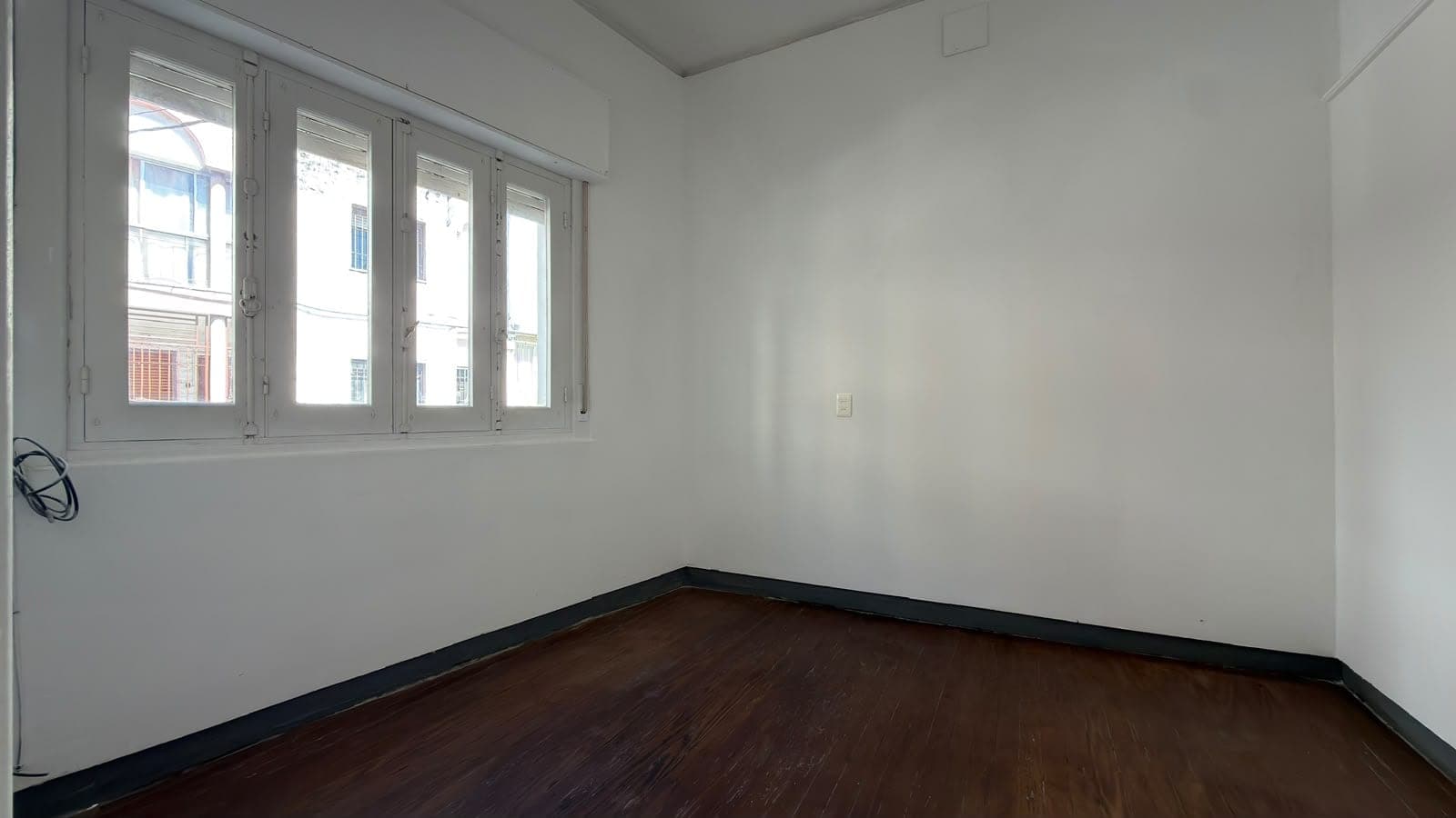 This is a bright, minimalist bedroom or living space featuring clean white walls and a dark wood floor with black baseboard trim. The room is flooded with natural light from a large multi-pane window system that offers views of neighboring buildings and street-level activity. The space appears empty and move-in ready, with simple finishes and a neutral aesthetic ideal for various uses.