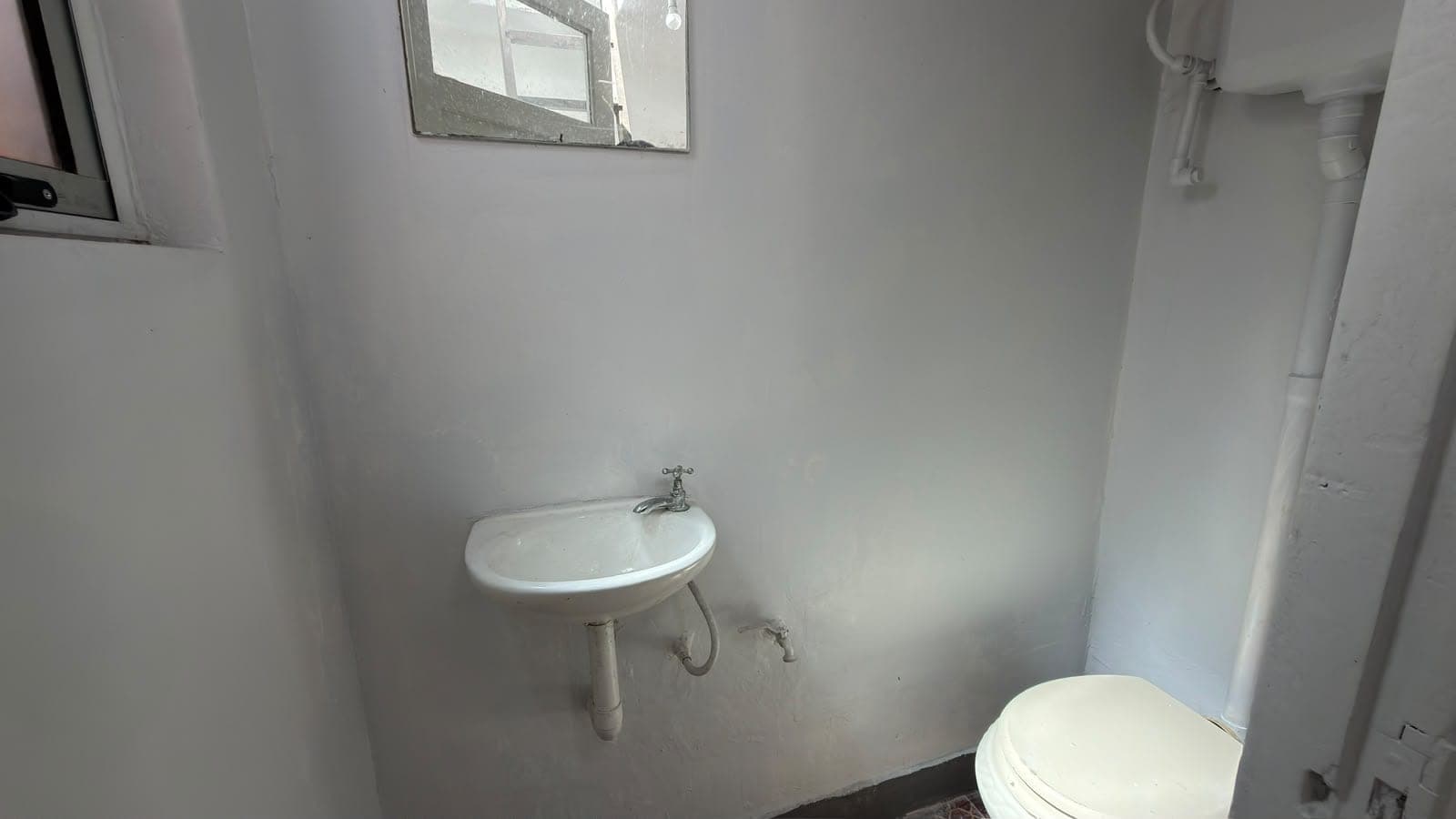 # Bathroom
This is a compact bathroom featuring basic fixtures including a wall-mounted round sink with chrome faucet and a toilet positioned in the lower right corner. The space has clean white painted walls, a small mirror above the sink, and a window providing natural light. The bathroom appears functional but minimal in finishes and would benefit from updates to fixtures and décor.