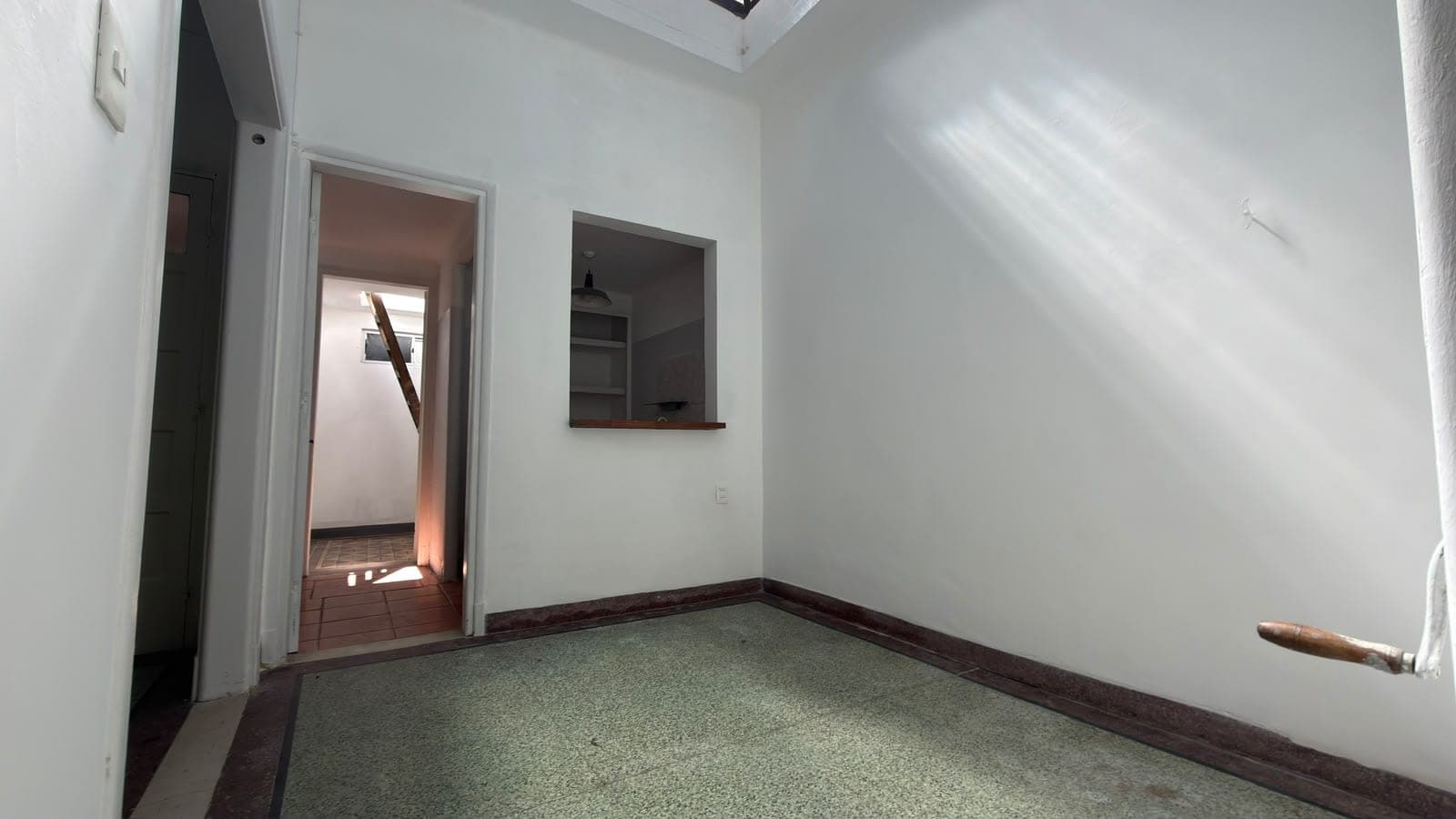 # Property Image Description
This is a bright, minimalist bedroom or living space featuring clean white walls and excellent natural light from a skylight above. The room includes green/gray carpeting with dark wood baseboards, a built-in wooden-framed window or pass-through opening to an adjacent room, and a dark wooden cabinet or shelving unit mounted on the wall. The space is well-maintained with neutral finishes and appears to be part of a multi-room residential unit with good flow between connected spaces.