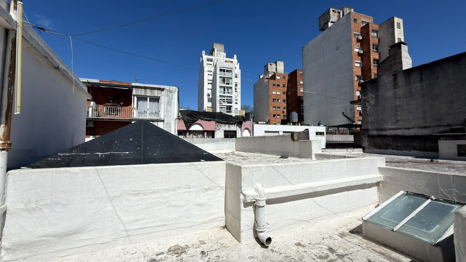 # Property Exterior - Rooftop/Terrace Space
This image shows a rooftop or terrace area of an urban residential building, featuring concrete flooring, structural elements, and utility infrastructure including skylights and drainage systems. The space is surrounded by multi-story apartment buildings in varying states of condition, with a clear blue sky overhead, indicating good weather and natural light exposure. The rooftop appears to be a common area or utility space with potential for development or renovation.