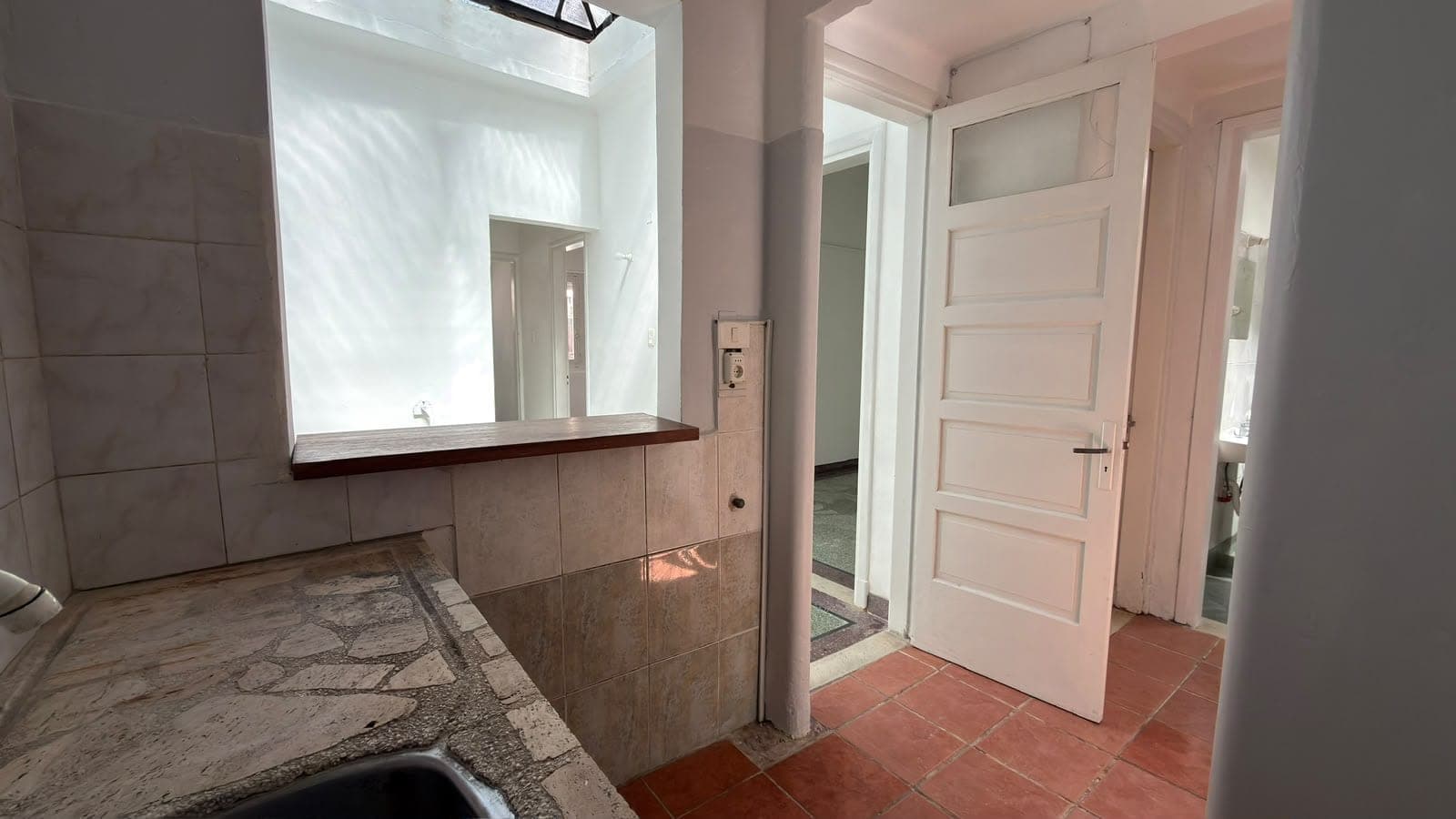 # Primary Bathroom
This spacious primary bathroom features a large vanity with a wooden-framed mirror and skylight above, providing excellent natural lighting. The space includes brown tile flooring, brown ceramic wall tiles, and multiple doorways providing access to adjoining rooms. The bathroom is currently in a pre-renovation or vacant state, showing good bones with solid fixtures and ample counter space.
