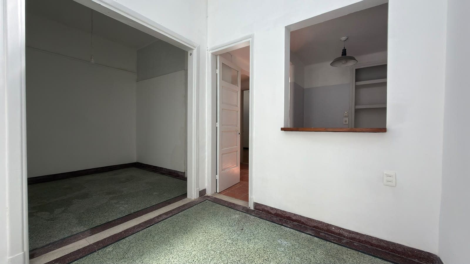 This is a spacious bedroom or master suite featuring clean white walls, terrazzo flooring with dark wood trim, and excellent natural light. The room includes a large walk-in closet on the left with mirrored doors, a wooden shelf unit on the right wall, and a black pendant light fixture, all in good condition. Additional features include visible electrical outlets and access to an adjoining room or hallway through a wooden door, making this a well-appointed residential space with modern finishes.